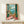 May include: Framed artwork displaying a Mediterranean balcony scene with green shutters, wicker chair, potted plants with pink flowers, and ocean view with distant mountains. The painting shows warm coral and turquoise tones with detailed interior elements like a small round table and decorative pottery. Artwork is positioned on a light wooden shelf next to a small wrapped gift with evergreen sprig.