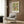 May include: Framed William Morris botanical poster with ornate floral pattern in greens, golds, and reds hanging on wood-paneled wall above cream upholstered armchair with geometric pillow. Modern brass floor lamp and dark round side table with open book complete the cozy reading nook scene. Natural lighting from window creates warm, sophisticated interior setting.