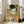 May include: Framed reproduction of Klimt's famous painting showing two embracing figures against ornate golden background with geometric patterns and colorful circular motifs. White wooden frame leans against bright living room wall beside beige upholstered chair near large windows with natural lighting on hardwood floors.