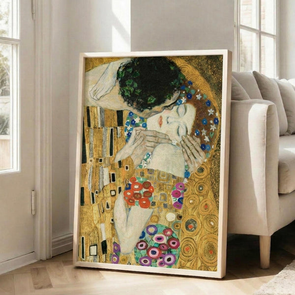 May include: Framed reproduction of Klimt's famous painting showing two embracing figures against ornate golden background with geometric patterns and colorful circular motifs. White wooden frame leans against bright living room wall beside beige upholstered chair near large windows with natural lighting on hardwood floors.