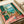 Mediterranean balcony scene with green shutters framing an ocean view. Wicker chair sits on coral pink tiled terrace with potted palm and flowering plants. Vintage travel poster style artwork in vibrant turquoise, coral, and green tones. Print displayed flat on white surface with neutral background and soft lighting.