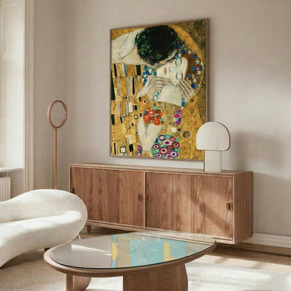 May include: Gustav Klimt's "The Kiss" art print displayed above a wooden sideboard in a modern living room. The iconic painting features two embracing figures in golden, ornate robes with geometric and floral patterns against a decorative background. The room shows a white curved chair, oval glass coffee table, floor lamp, and neutral walls with natural lighting.