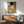 May include: Gustav Klimt's "The Kiss" reproduction poster displayed in modern living room setting with wooden credenza, sage green mid-century armchair, round coffee table, and white ceramic lamp. The iconic Art Nouveau painting features two embracing figures in ornate golden robes with geometric and floral patterns against a bronze background, shown in a thin wooden frame on white wall with natural sunlight streaming across the scene.