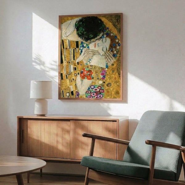 May include: Gustav Klimt's "The Kiss" reproduction poster displayed in modern living room setting with wooden credenza, sage green mid-century armchair, round coffee table, and white ceramic lamp. The iconic Art Nouveau painting features two embracing figures in ornate golden robes with geometric and floral patterns against a bronze background, shown in a thin wooden frame on white wall with natural sunlight streaming across the scene.