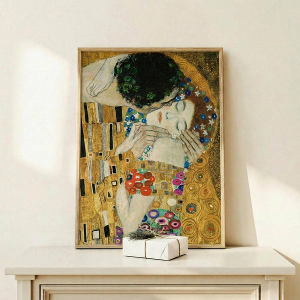 May include: Framed reproduction of Klimt's famous painting showing two embracing figures against ornate gold patterned background with geometric and circular motifs in yellow, green, blue, and red. The woman has dark hair and closed eyes while decorative robes flow around both figures. Print displayed on white shelf with small wrapped gift beside it.