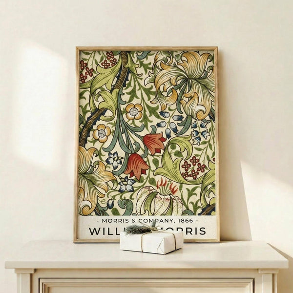 May include: Framed botanical poster with intricate floral pattern featuring green leaves, golden flowers, red berries, and ornate scrollwork in William Morris Victorian decorative style, displayed on light wood shelf next to small white wrapped gift with evergreen sprig against cream colored wall