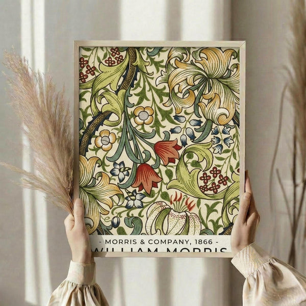 May include: Hands holding a framed botanical art print featuring William Morris-style decorative pattern with flowing vines, colorful flowers in yellow, red and blue, ornate leaves, and berries on cream background. Person wearing beige ruffled sleeves holds dried pampas grass stems against neutral curtained backdrop.