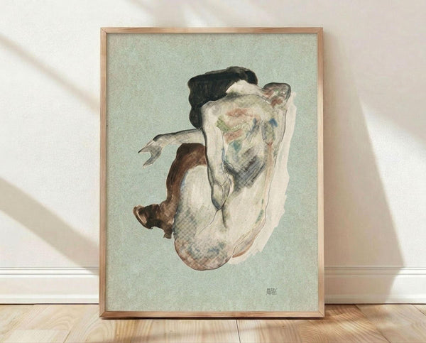May include: Framed reproduction of an expressionist watercolor drawing showing a crouching female figure with dark hair against a pale green background. The artwork features loose, gestural brushstrokes in muted tones of brown, pink, and blue. The thin wooden frame sits on light wooden flooring against a white textured wall with soft natural lighting casting diagonal shadows across the scene.