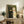 May include: Framed vintage-style painting of two cats in formal wedding attire displayed on rustic wooden sideboard. Black cat wears yellow bow tie, white cat wears pink bow and holds yellow bouquet. Green background, light wood frame. Room scene shows beige chair, fern plant, pottery, and books creating cozy home decor setting with neutral wall.