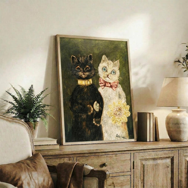 May include: Framed vintage-style painting of two cats in formal wedding attire displayed on rustic wooden sideboard. Black cat wears yellow bow tie, white cat wears pink bow and holds yellow bouquet. Green background, light wood frame. Room scene shows beige chair, fern plant, pottery, and books creating cozy home decor setting with neutral wall.