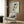 May include: Framed art print of a crouching female figure in earth tones and pale green, displayed on wood-paneled wall above a cream upholstered armchair with geometric pillow. Modern reading nook features brass floor lamp and dark round side table with open book.