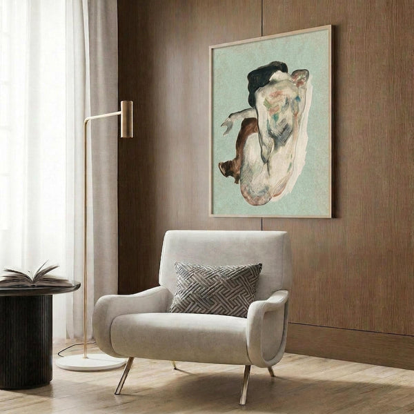 May include: Framed art print of a crouching female figure in earth tones and pale green, displayed on wood-paneled wall above a cream upholstered armchair with geometric pillow. Modern reading nook features brass floor lamp and dark round side table with open book.