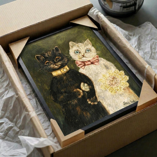 May include: Vintage-style illustration of two cats in ornate dress - a black cat wearing a golden collar and a white cat with blue eyes wearing a pink bow and yellow lace dress. The artwork has a dark olive green background and appears to be a framed print being carefully unpacked from a cardboard shipping box with white tissue paper protection.