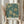 May include: Hands holding a framed art print featuring an intricate botanical pattern in teal and cream colors. The design shows flowing floral motifs with chrysanthemums, leaves, and ornate scrollwork in William Morris's distinctive Arts and Crafts style. The print has a light wooden frame and includes text reading "Morris & Company, 1866" and "William Morris" at the bottom. Dried pampas grass stems are visible to the left, and soft beige curtains create a neutral backdrop.