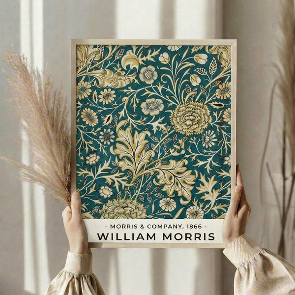 May include: Hands holding a framed art print featuring an intricate botanical pattern in teal and cream colors. The design shows flowing floral motifs with chrysanthemums, leaves, and ornate scrollwork in William Morris's distinctive Arts and Crafts style. The print has a light wooden frame and includes text reading "Morris & Company, 1866" and "William Morris" at the bottom. Dried pampas grass stems are visible to the left, and soft beige curtains create a neutral backdrop.