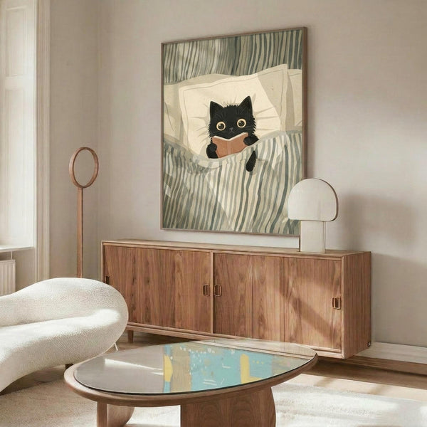 May include: Whimsical illustration of a black cat with large yellow eyes reading a small brown book while tucked under green and white striped bedding. The artwork is displayed in a wooden frame above a walnut credenza in a mid-century modern living room with cream walls, white upholstered chair, glass coffee table, and warm natural lighting.