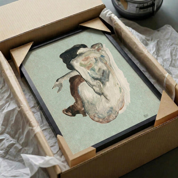 Framed art print of a crouching nude woman with dark hair against a pale green background, shown in expressionist style with earth tones and loose brushwork. The black-framed artwork is being unpacked from a cardboard box with protective tissue paper and wooden corner guards, photographed on a white surface next to what appears to be a dark glass object.