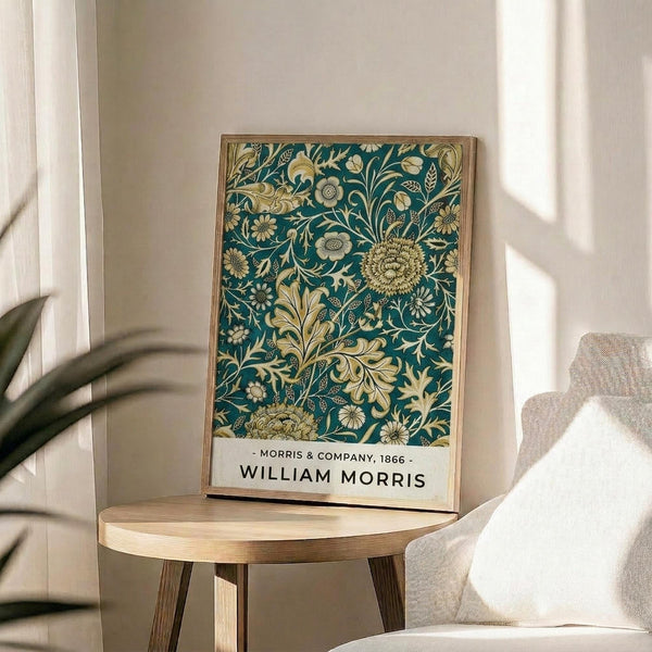 May include: Framed William Morris botanical poster with teal blue background featuring ornate gold and cream floral pattern including chrysanthemums, leaves, and decorative vines. Light wood frame sits on round wooden side table next to beige upholstered chair. Soft natural lighting creates shadows on white wall with plant visible in corner of minimalist room setting.