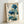 May include: Framed botanical art print featuring blue poppies with delicate petals and seed pods, accompanied by traditional Asian calligraphy text on aged cream paper background. The artwork is displayed in a simple light wood frame, leaning against a white wall with soft natural lighting creating gentle shadows.