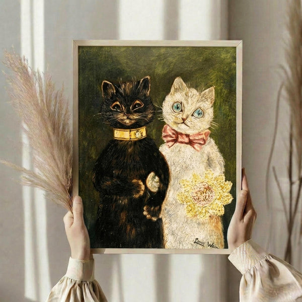 May include: Person holding framed vintage-style cat wedding artwork featuring black cat in golden collar and white cat in pink bow tie holding yellow bouquet against green background, displayed in light wood frame in bright interior with dried pampas grass and white curtains