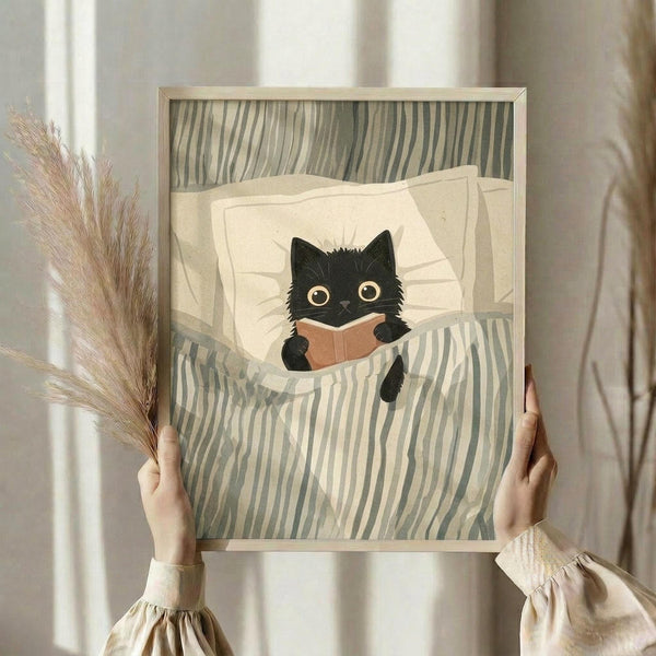 May include: Hands holding a framed art print showing a black cat with large golden eyes reading a small brown book while tucked in bed under green and cream striped bedding. The illustration has a cozy, whimsical style with soft colors. Person wearing cream-colored clothing with gathered sleeves holds dried pampas grass in one hand. Neutral bedroom setting with white curtains and warm natural lighting.