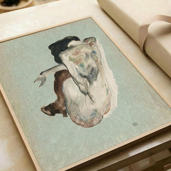 May include: Framed reproduction of an early 20th century expressionist drawing showing a crouching nude female figure with dark hair against a pale green background. The artwork features loose, gestural lines in earth tones and muted colors. The print is displayed in a light wood frame on white paper, positioned on a cream-colored surface next to neutral fabric and ribbon in soft lighting.