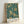 A framed poster featuring an intricate botanical pattern on a teal background with cream and golden yellow flowers, leaves, and decorative flourishes in Victorian Arts and Crafts style. The design includes detailed chrysanthemums, ornate foliage, and flowing organic motifs. Text at bottom reads "Morris & Company, 1866 - William Morris" on a cream banner. The poster is displayed in a light wooden frame, leaning against a white wall with soft natural lighting creating gentle shadows.