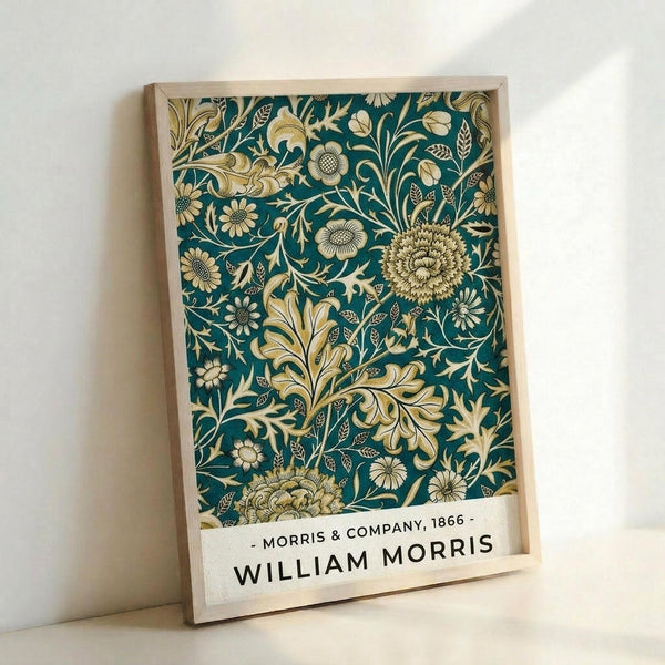 A framed poster featuring an intricate botanical pattern on a teal background with cream and golden yellow flowers, leaves, and decorative flourishes in Victorian Arts and Crafts style. The design includes detailed chrysanthemums, ornate foliage, and flowing organic motifs. Text at bottom reads "Morris & Company, 1866 - William Morris" on a cream banner. The poster is displayed in a light wooden frame, leaning against a white wall with soft natural lighting creating gentle shadows.