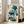 May include: Framed botanical artwork featuring blue poppies with Asian calligraphy text, displayed on wooden floor beside beige upholstered chair near white-framed windows. The print shows detailed blue flowers with green stems and seed pods against cream background, housed in light wood frame. Natural sunlight streams across white walls and light hardwood flooring in modern living room setting.