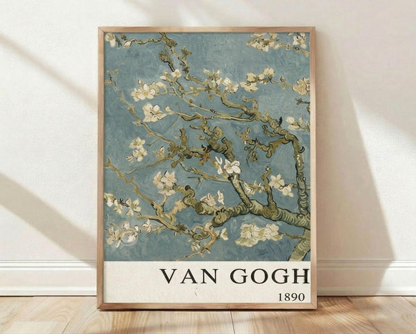 May include: Framed poster print of Van Gogh's almond blossom painting featuring white flowering branches against soft blue background, displayed in thin wood frame leaning against white wall on light wooden floor. Delicate brushstrokes show gnarled brown branches with clusters of pale pink and white blossoms scattered throughout the composition.