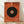 May include: Framed abstract art print leaning against white wall on light wood floor. Orange textured background with large black circle containing concentric rings in orange, blue, and yellow at center. Two diagonal lines extend from circle - one yellow, one purple. Minimalist geometric composition in thin light wood frame.