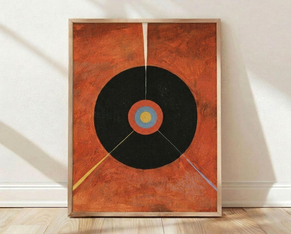 May include: Framed abstract art print leaning against white wall on light wood floor. Orange textured background with large black circle containing concentric rings in orange, blue, and yellow at center. Two diagonal lines extend from circle - one yellow, one purple. Minimalist geometric composition in thin light wood frame.
