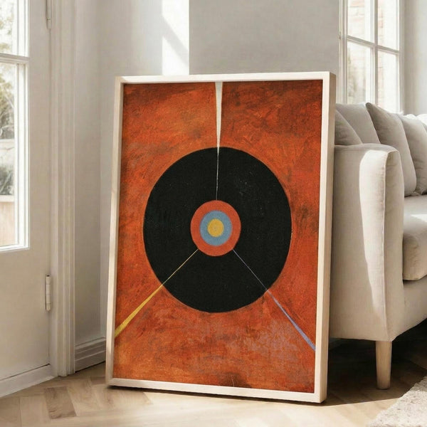 May include: Large framed abstract art print leaning against white wall next to beige sofa in bright living room. Orange textured background with large black circle containing smaller concentric circles in orange, blue and yellow. Thin colored lines extend from center like rays. Natural light from tall windows creates soft shadows on hardwood floor.