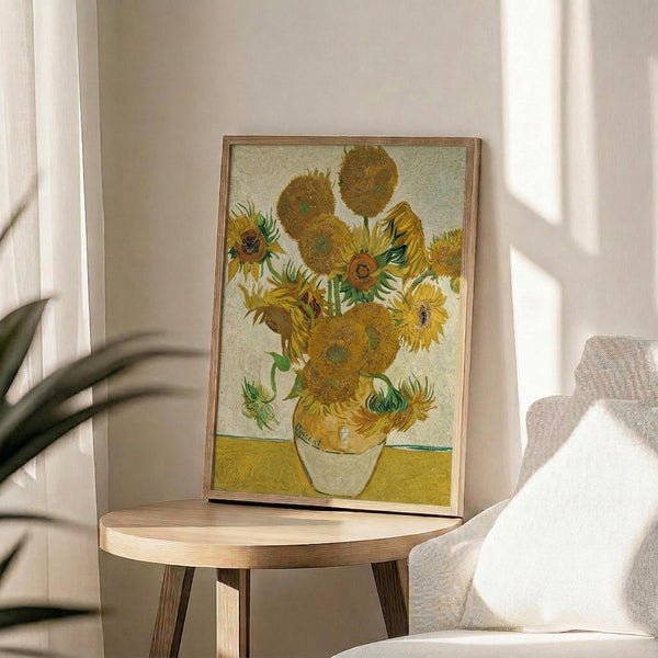 May include: Van Gogh's famous sunflowers painting reproduction in thin wooden frame, displayed on round wooden side table next to beige upholstered chair. Artwork shows vibrant yellow sunflowers in white vase against yellow surface and pale background. Warm natural lighting creates soft shadows across neutral-toned room with plant visible in corner.