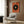 May include: Abstract geometric art print with orange background featuring large black circle with blue and yellow center, displayed in light wooden frame on wood-paneled wall. Modern beige armchair with patterned pillow, brass floor lamp, and dark round side table with open book visible in contemporary interior setting.
