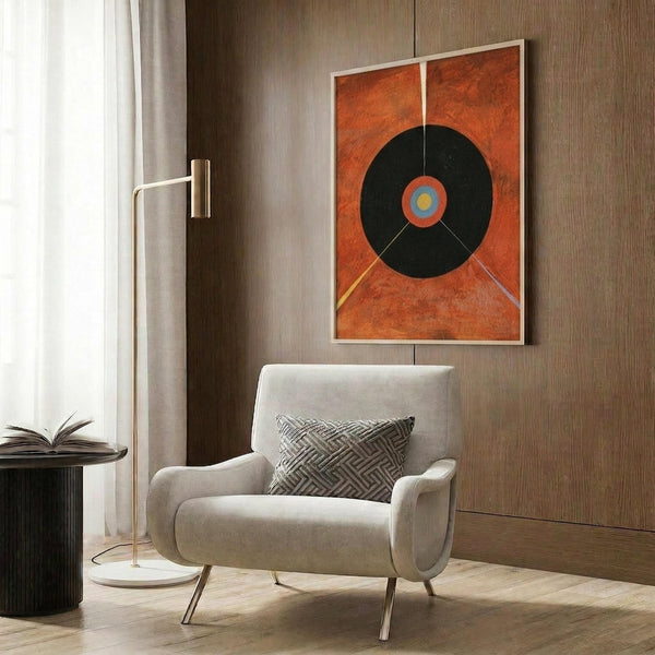May include: Abstract geometric art print with orange background featuring large black circle with blue and yellow center, displayed in light wooden frame on wood-paneled wall. Modern beige armchair with patterned pillow, brass floor lamp, and dark round side table with open book visible in contemporary interior setting.