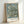 May include: Framed poster print leaning against white wall showing Van Gogh's almond blossom painting with delicate white flowers on twisted brown branches against soft blue-green background. Light wood frame with white border containing "VAN GOGH 1890" text below artwork. Natural lighting creates gentle shadows on minimalist white surface.