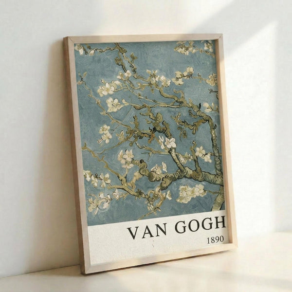 May include: Framed poster print leaning against white wall showing Van Gogh's almond blossom painting with delicate white flowers on twisted brown branches against soft blue-green background. Light wood frame with white border containing "VAN GOGH 1890" text below artwork. Natural lighting creates gentle shadows on minimalist white surface.