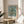 May include: Framed poster featuring Van Gogh's almond blossom painting with white flowers on turquoise blue background, displayed on round wooden side table next to beige upholstered chair. Natural sunlight streams across white walls, with green plant leaves visible in corner. Poster has thin wooden frame and museum-style text below artwork.