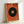 Framed abstract poster featuring a large black circle on textured orange background, with concentric rings of orange, blue and yellow at center, and thin colored lines extending from the circle. The artwork is displayed in a light wood frame leaning against a white wall.