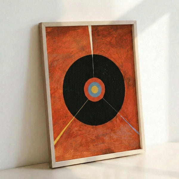 Framed abstract poster featuring a large black circle on textured orange background, with concentric rings of orange, blue and yellow at center, and thin colored lines extending from the circle. The artwork is displayed in a light wood frame leaning against a white wall.