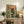May include: Framed poster artwork displayed on a rustic wooden sideboard in a cozy living room setting. The illustration features a black cat wearing pink glasses peering over a large glass fishbowl containing orange goldfish and aquatic plants, surrounded by coral-pink background with green botanical elements. Scene includes fern plant, books, ceramic vase, and beige upholstered furniture with brown throw pillows.