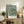 May include: Van Gogh's Almond Blossom artwork featuring white flowering branches against a soft blue background, displayed in a light wooden frame on a rustic wooden sideboard. The poster shows delicate almond blossoms with twisted brown branches in Van Gogh's distinctive style. Scene includes a cream upholstered chair, potted fern plant, stacked books, and ceramic vase creating a warm, natural living space setting.
