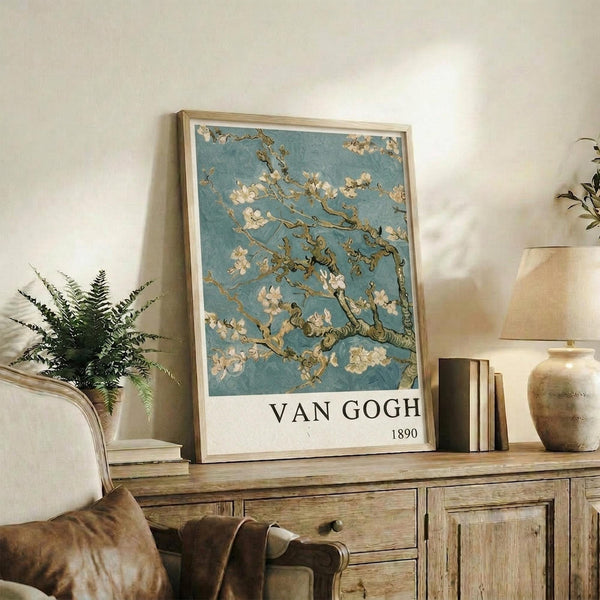 May include: Van Gogh's Almond Blossom artwork featuring white flowering branches against a soft blue background, displayed in a light wooden frame on a rustic wooden sideboard. The poster shows delicate almond blossoms with twisted brown branches in Van Gogh's distinctive style. Scene includes a cream upholstered chair, potted fern plant, stacked books, and ceramic vase creating a warm, natural living space setting.