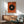 May include: Framed abstract art print with large black circle on orange textured background, featuring colorful center detail and geometric lines, displayed above wooden sideboard with white table lamp in modern living room setting with sage green upholstered chair and natural lighting creating dramatic shadows on white wall.