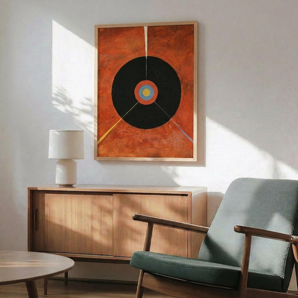 May include: Framed abstract art print with large black circle on orange textured background, featuring colorful center detail and geometric lines, displayed above wooden sideboard with white table lamp in modern living room setting with sage green upholstered chair and natural lighting creating dramatic shadows on white wall.