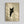May include: Framed poster print hanging on white wall showing black cat with golden yellow eyes peeking around corner or doorway. Artwork features minimalist beige and cream color palette with geometric shadow patterns. Cat illustration shows detailed whiskers and alert expression. Natural wood frame with simple border. Soft lighting creates diagonal shadows across the wall surface.
