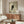 May include: Black cat portrait with bright yellow eyes peeking from corner, displayed in thin wooden frame above walnut credenza in modern living room. Artwork shows minimalist illustration on sage green background. Room features cream upholstered chair, glass-top coffee table, white table lamp, and natural window light casting geometric shadows across walls and furniture.