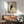 May include: A framed poster print of a black cat with bright yellow eyes peeking from behind a light background, displayed on a white wall above a wooden sideboard. The minimalist artwork features clean lines and is shown in a modern living room setting with a mint green armchair, small table lamp, and natural sunlight casting geometric shadows across the scene.