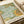 Decorative botanical print featuring intricate William Morris-style design with flowing acanthus leaves in sage green, golden yellow flowers, and ornate curved stems on a turquoise background. The vintage-inspired artwork displays fine line work and organic Art Nouveau patterns. Print shown flat on white paper with cream ribbon visible at edge, demonstrating the poster's size and paper texture.